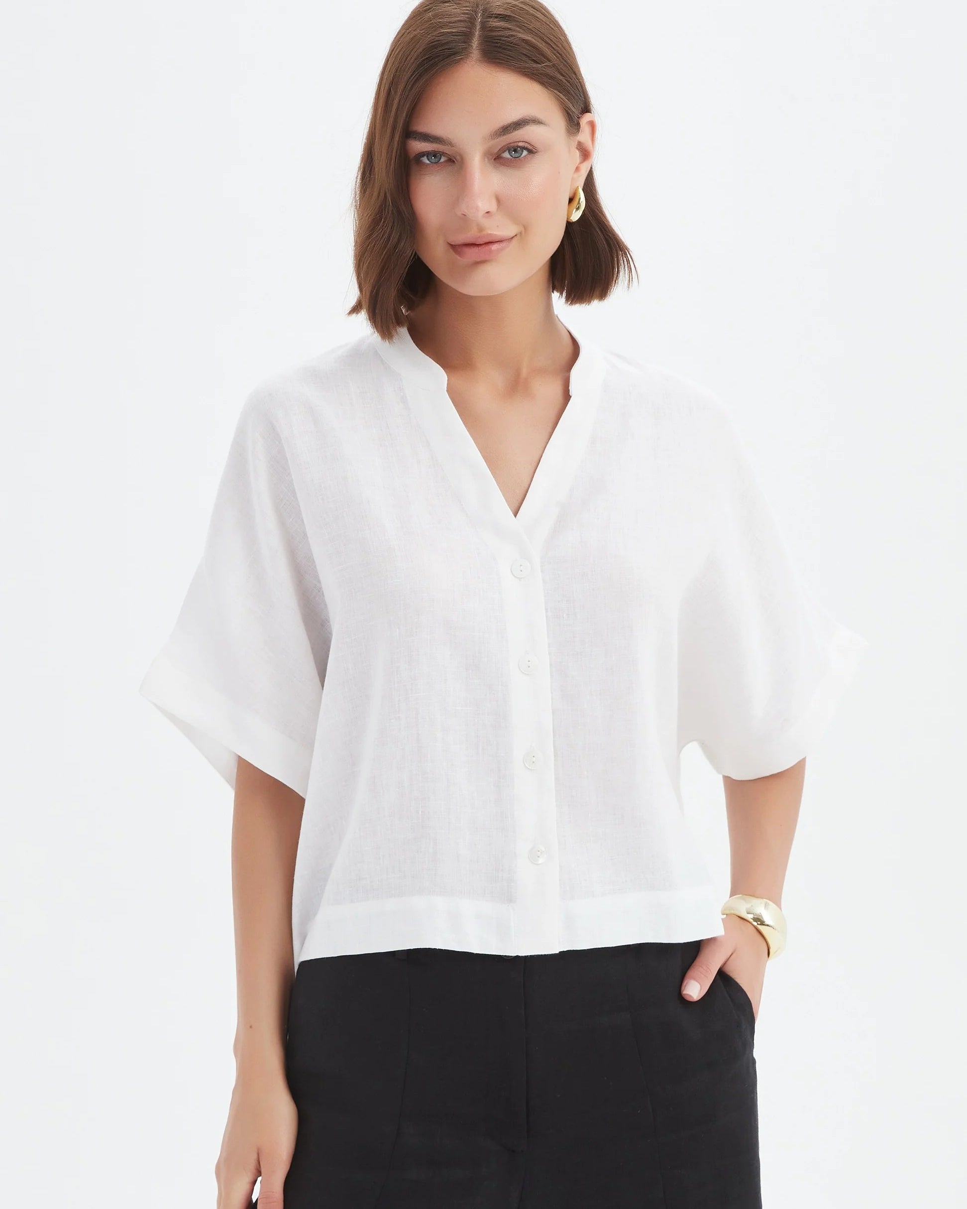 A woman modeling a white short-sleeve linen blouse with a split V-neck and black trousers, photographed against a clean white studio background.