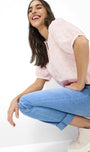 A woman crouching in a studio setting, wearing a light pink textured short-sleeve blouse, light blue jeans, and pale pink platform sneakers against a plain white background.