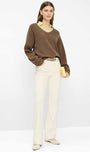 A model stands against a clean white studio background wearing a brown knit sweater layered over a pale yellow collared shirt, cream-colored trousers, and beige flats, while holding a small yellow clutch.