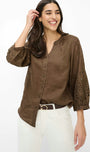 A woman modeling a brown button-up blouse with eyelet lace sleeves, paired with white pants and a brown belt, against a plain white background.