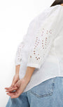 A woman wearing a white eyelet blouse with three-quarter sleeves and light blue jeans, shown from a side-back angle against a plain white background.