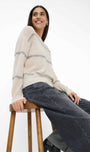 Woman sitting on a wooden stool wearing a cream sweater with blue stripes and dark jeans.