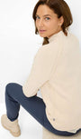 Woman wearing a beige sweater and dark blue pants sitting on a white surface.