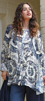 A woman wearing a long sleeve button up tunic top with a blue and white paisley pattern, paired with dark blue pants, standing outdoors against a cream coloured brick wall.