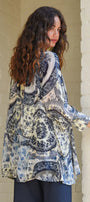 A woman with long, curly brown hair is shown from the back wearing a loose, long sleeve patterned blouse with a blue, grey, and cream paisley design, paired with dark pants, standing against a light coloured brick wall.