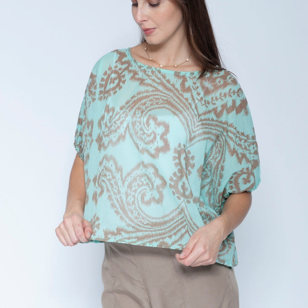 A woman wearing a light mint green short sleeve blouse with a brown paisley pattern, paired with beige trousers, standing against a light gray studio background.