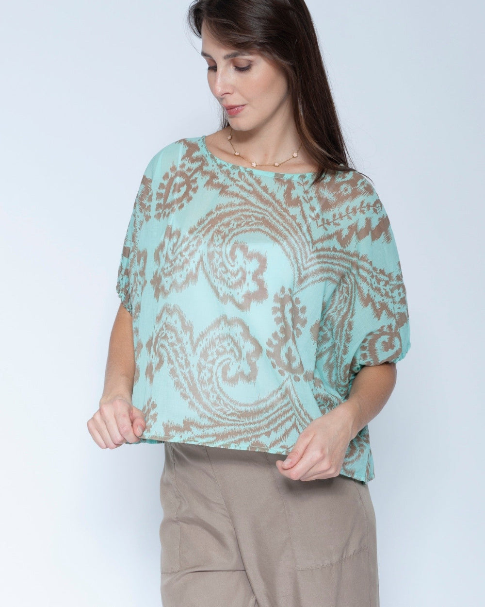 A woman wearing a light mint green short sleeve blouse with a brown paisley pattern, paired with beige trousers, standing against a light gray studio background.