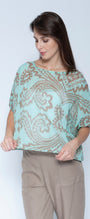 A woman wearing a light mint green short sleeve blouse with a brown paisley pattern, paired with beige trousers, standing against a light gray studio background.