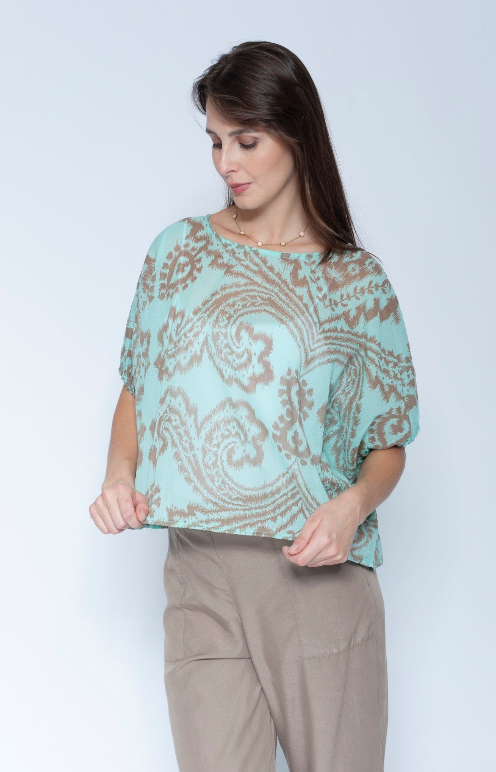 A woman wearing a light mint green short sleeve blouse with a brown paisley pattern, paired with beige trousers, standing against a light gray studio background.