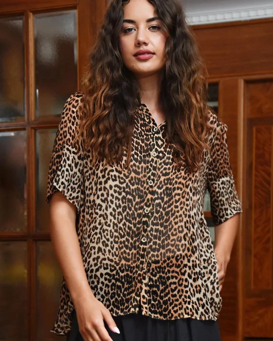 A woman with long, wavy brown hair wearing a leopard print short sleeve blouse and a black skirt, standing in front of a wooden cabinet.