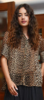 A woman with long, wavy brown hair wearing a leopard print short sleeve blouse and a black skirt, standing in front of a wooden cabinet.