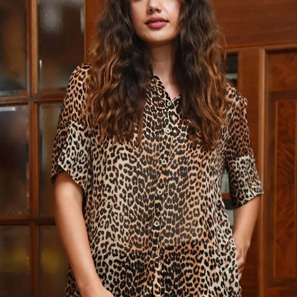 A woman with long, wavy brown hair wearing a leopard print short sleeve blouse and a black skirt, standing in front of a wooden cabinet.