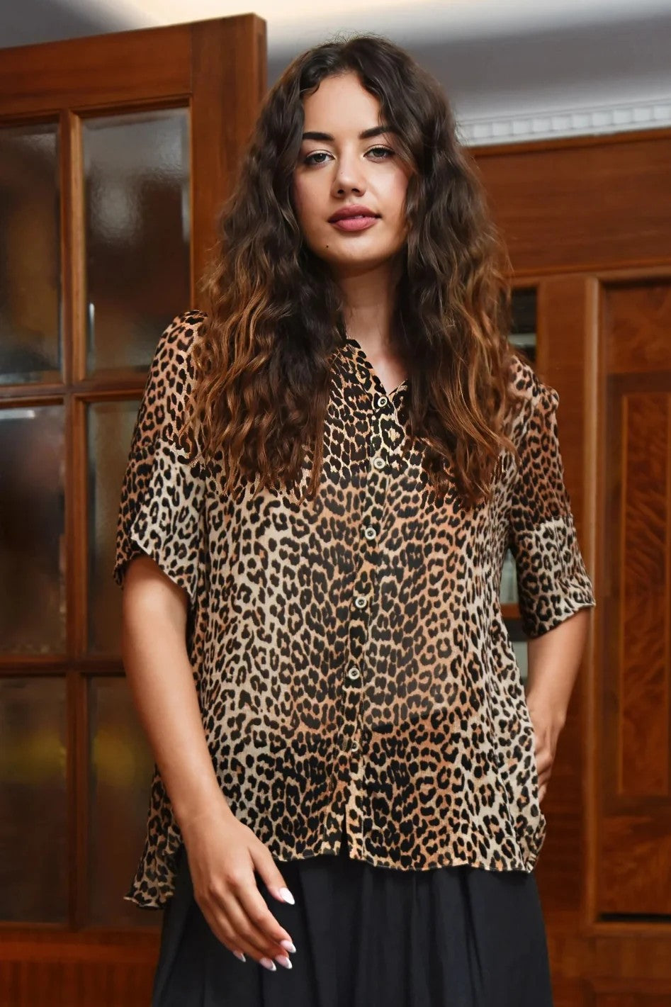 A woman with long, wavy brown hair wearing a leopard print short sleeve blouse and a black skirt, standing in front of a wooden cabinet.
