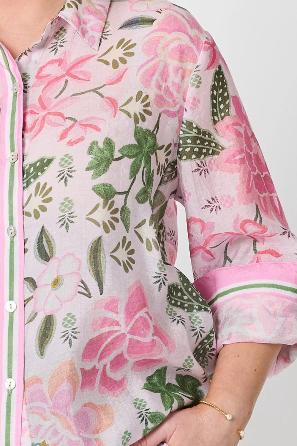 A close up of a woman wearing a light pink button up shirt with a large floral print in pink and green, featuring rolled up sleeves 