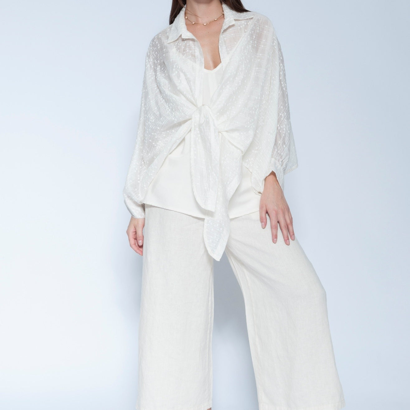 A woman modeling a matching off white loungewear set consisting of a long sleeve tie front shirt and wide leg cropped pants, photographed against a light gray studio background.