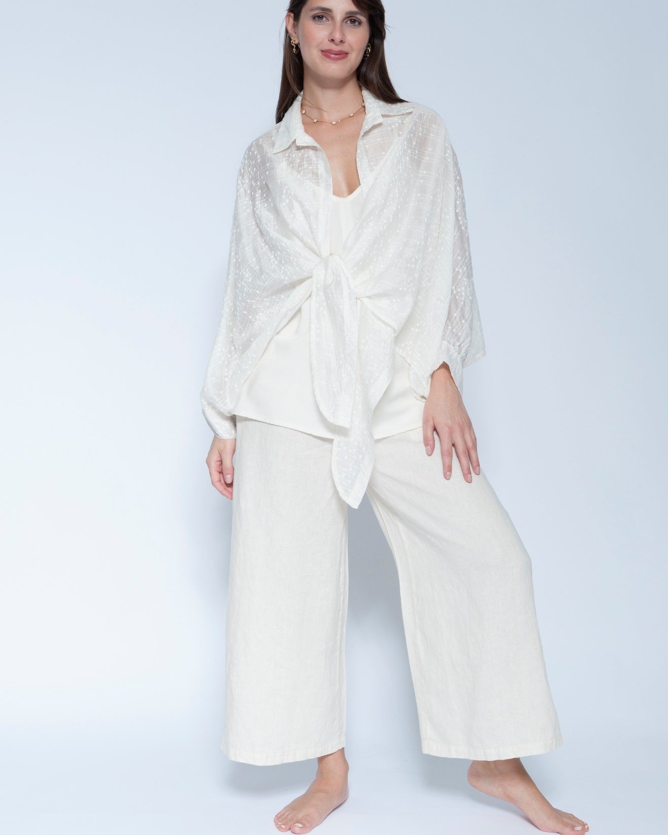 A woman modeling a matching off white loungewear set consisting of a long sleeve tie front shirt and wide leg cropped pants, photographed against a light gray studio background.