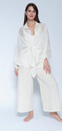A woman modeling a matching off white loungewear set consisting of a long sleeve tie front shirt and wide leg cropped pants, photographed against a light gray studio background.