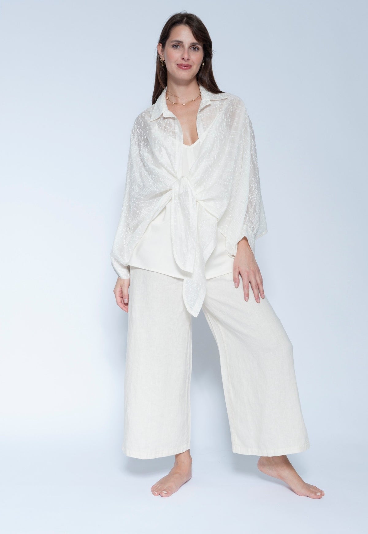 A woman modeling a matching off white loungewear set consisting of a long sleeve tie front shirt and wide leg cropped pants, photographed against a light gray studio background.