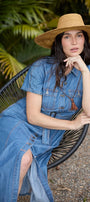 A woman wearing a blue denim dress and a straw hat sits in a black woven chair outdoors in front of lush green foliage.