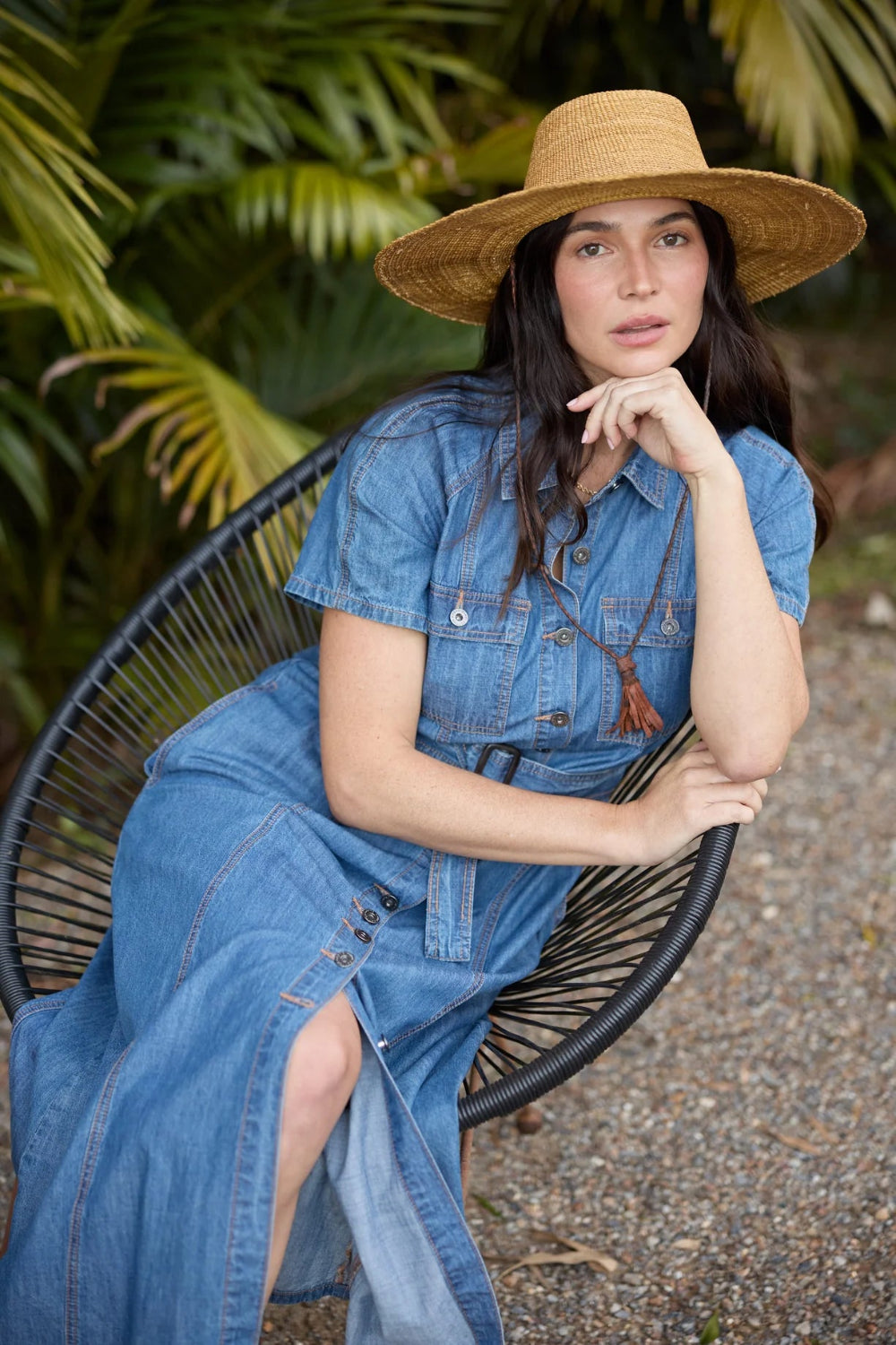 A woman wearing a blue denim dress and a straw hat sits in a black woven chair outdoors in front of lush green foliage.