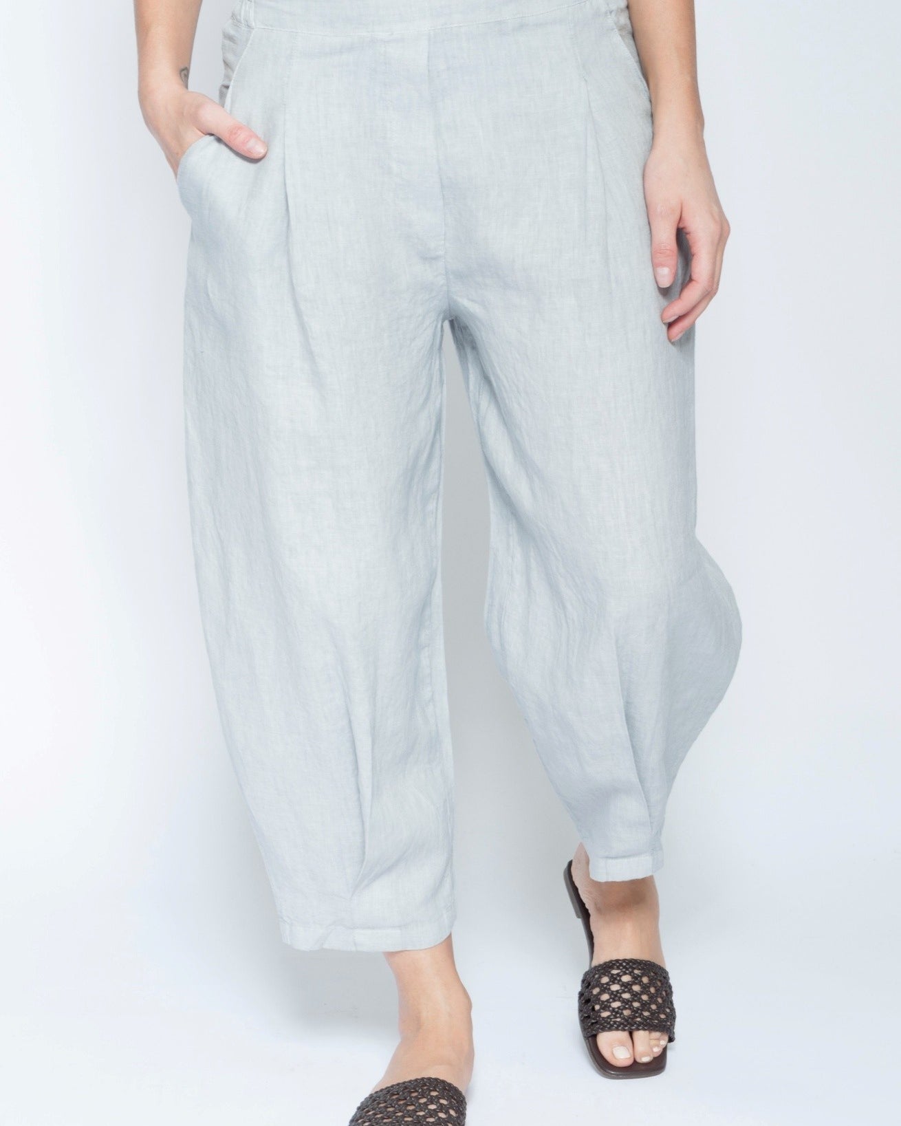 A person wearing light gray wide-leg cropped pants with side pockets, paired with black woven slide sandals, shown against a plain white studio background.