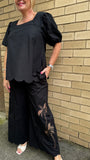 Person wearing a black pant with floral embroidery against a tiled wall.