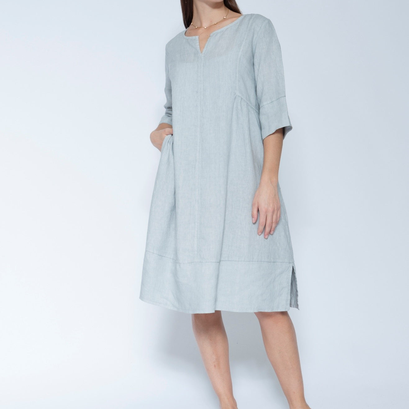 A woman modleling a light blue grey linen tunic dress with three quarter sleeves and a split neckline, paired with white slide sandals.
