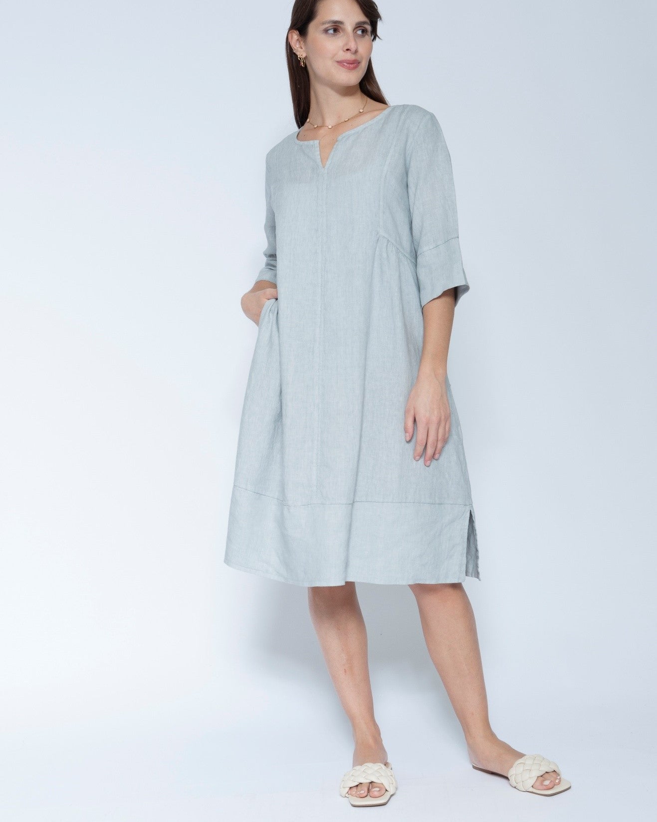 A woman modleling a light blue grey linen tunic dress with three quarter sleeves and a split neckline, paired with white slide sandals.