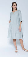 A woman modleling a light blue grey linen tunic dress with three quarter sleeves and a split neckline, paired with white slide sandals.