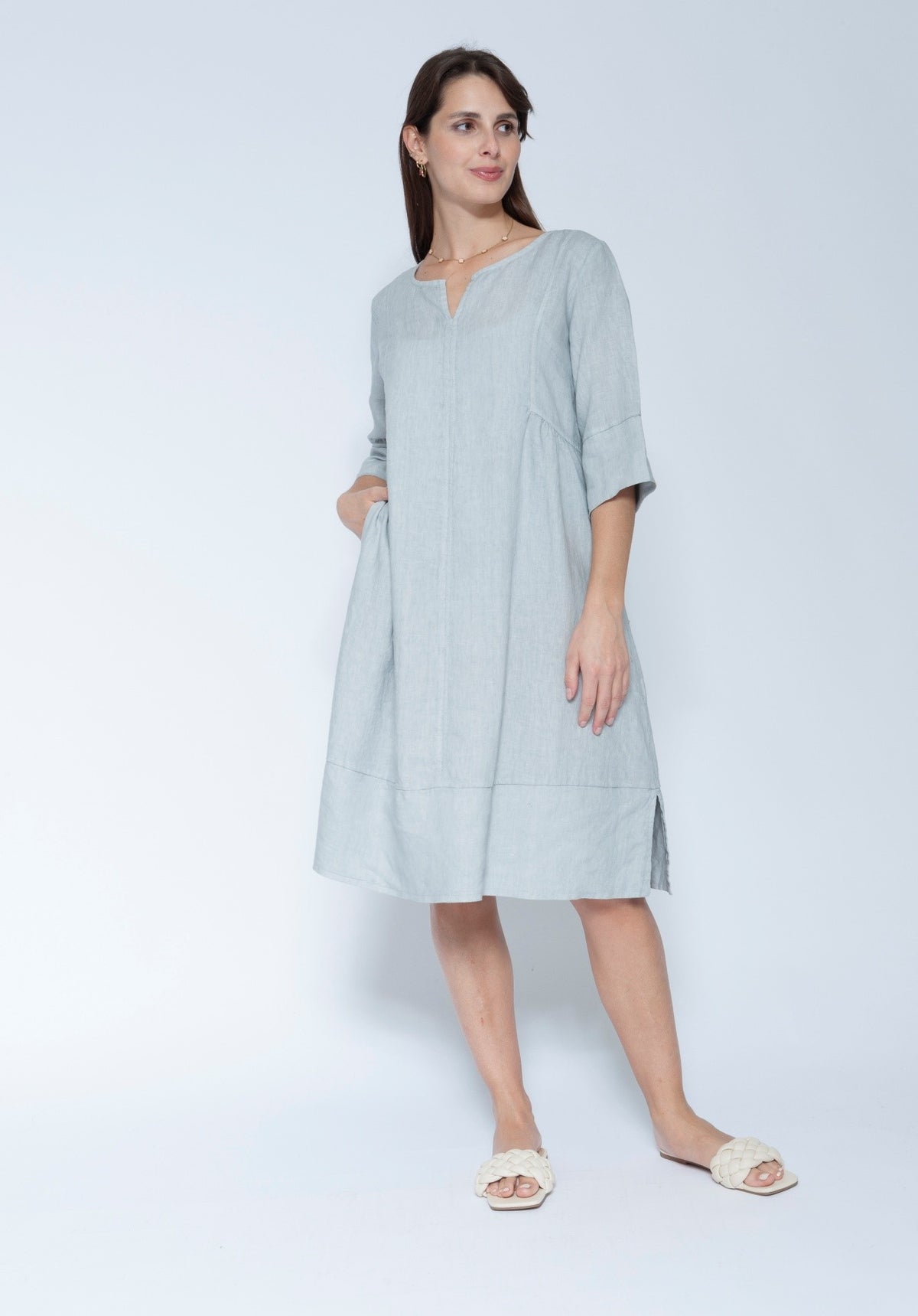 A woman modleling a light blue grey linen tunic dress with three quarter sleeves and a split neckline, paired with white slide sandals.