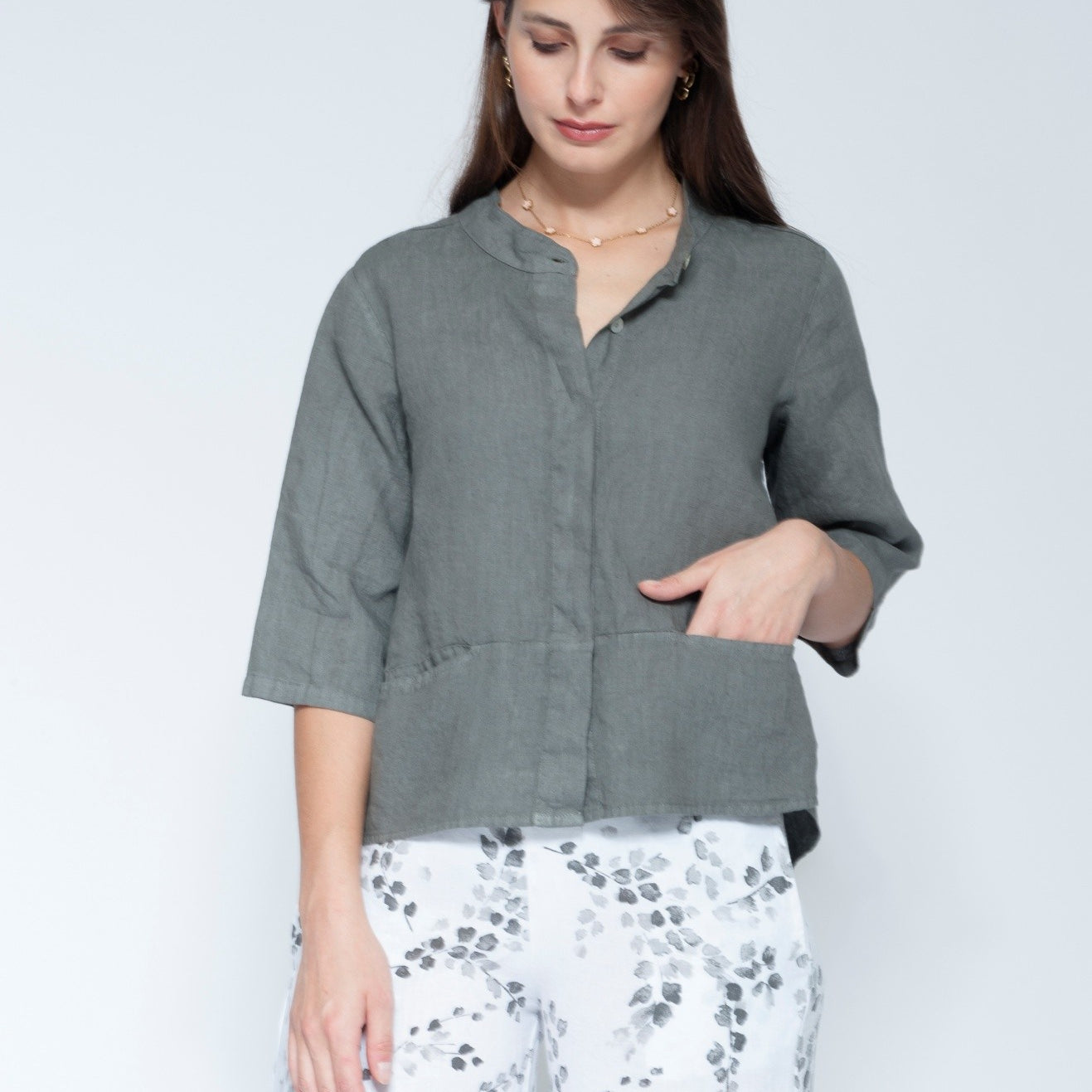 Front view muted grey green linen button up shirt with three quarter sleeves and front pockets, paired with white pants featuring a grey floral pattern.