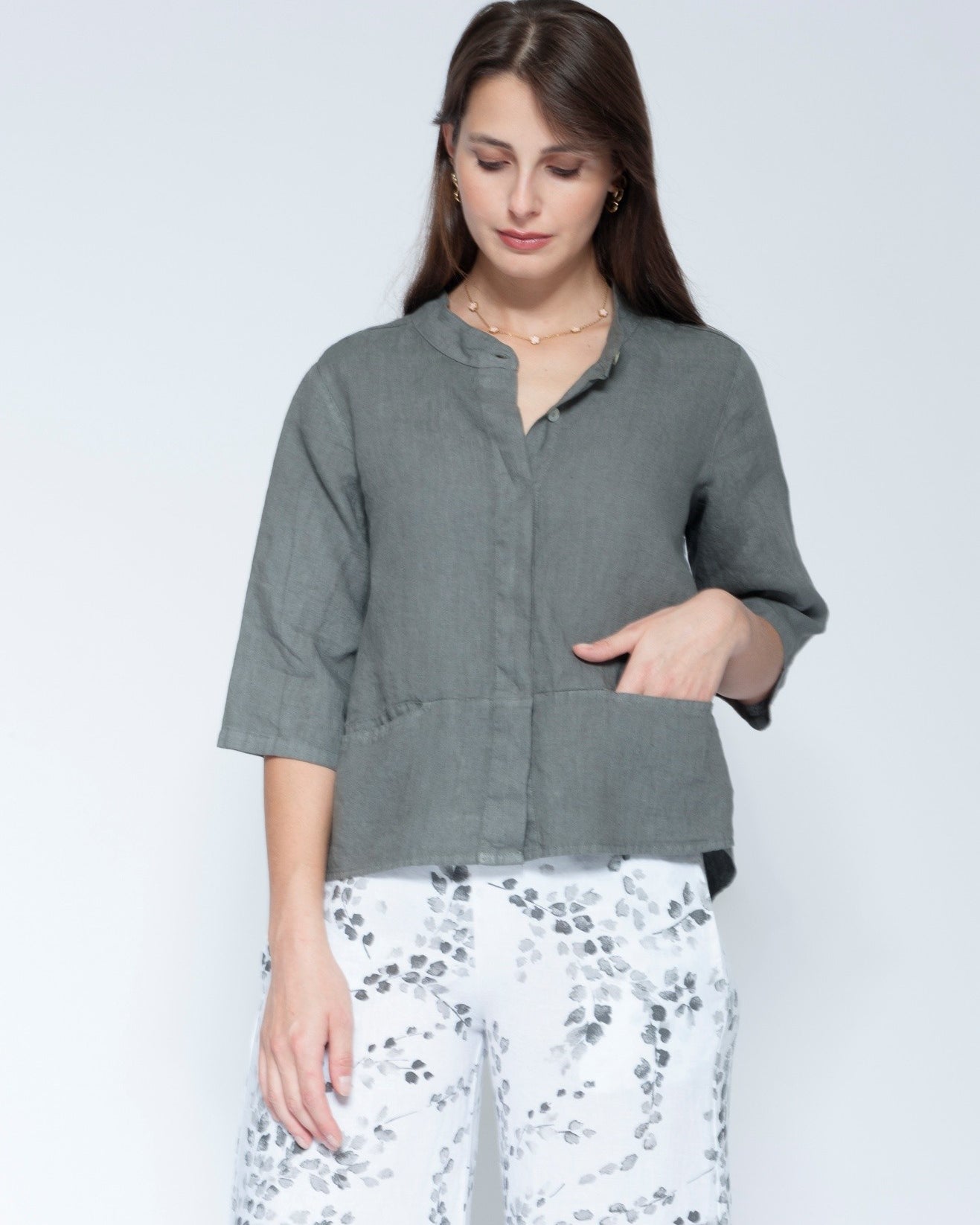 Front view muted grey green linen button up shirt with three quarter sleeves and front pockets, paired with white pants featuring a grey floral pattern.