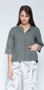 Front view muted grey green linen button up shirt with three quarter sleeves and front pockets, paired with white pants featuring a grey floral pattern.