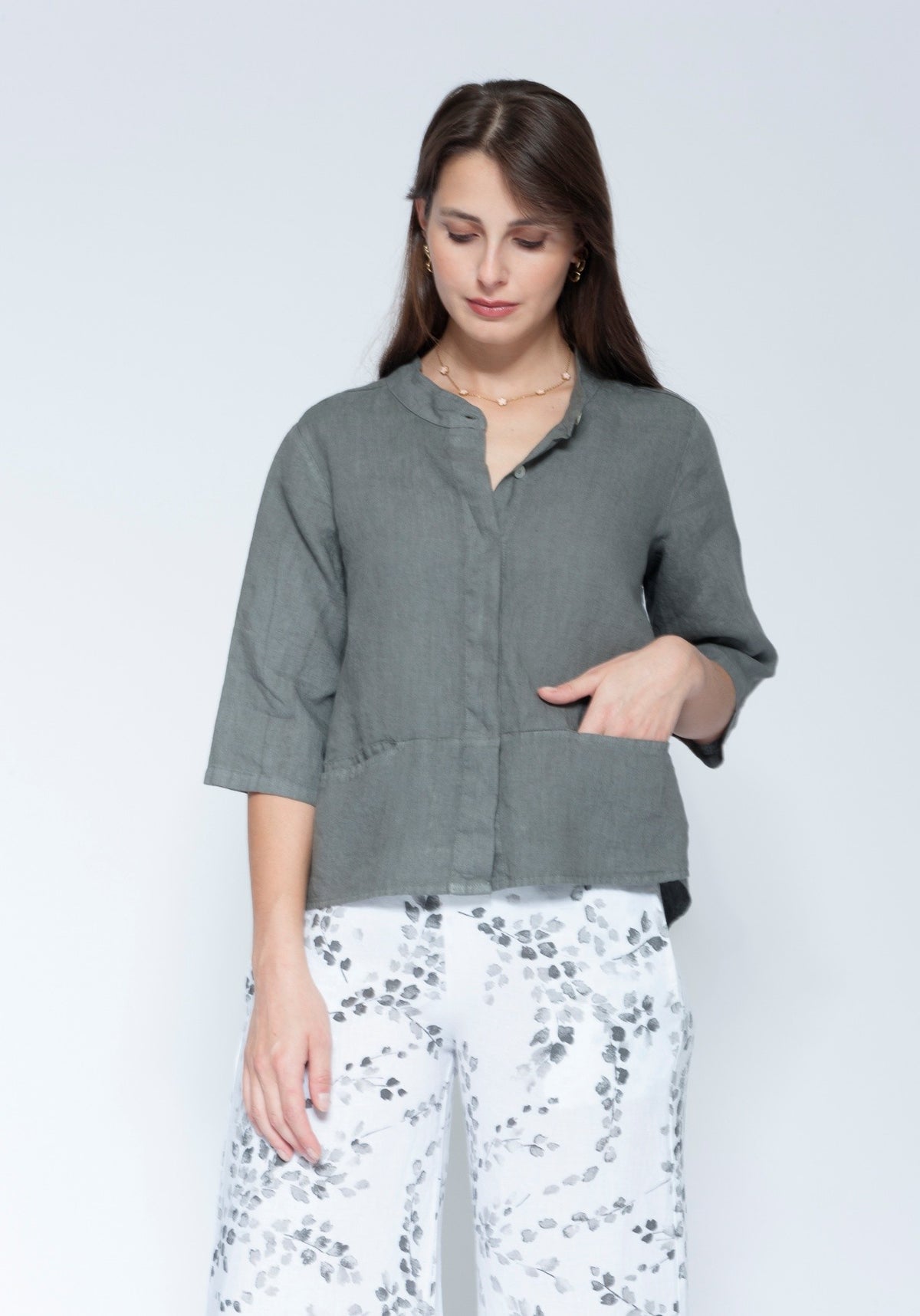 Front view muted grey green linen button up shirt with three quarter sleeves and front pockets, paired with white pants featuring a grey floral pattern.
