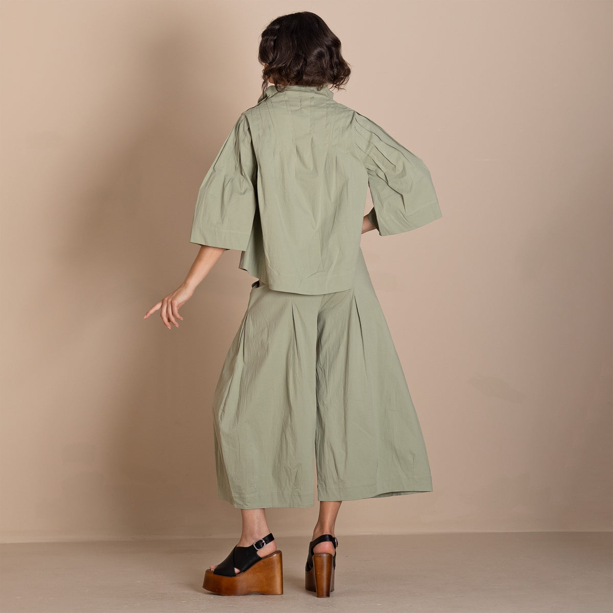 Back view a sage green two piece outfit consisting of a short sleeve cropped shirt and matching wide leg cropped pants, paired with black wedge sandals.