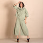A woman models a light sage green two piece outfit consisting of a short sleeve, high neck, ruffled top and matching wide leg cropped pants, paired with black platform sandals.