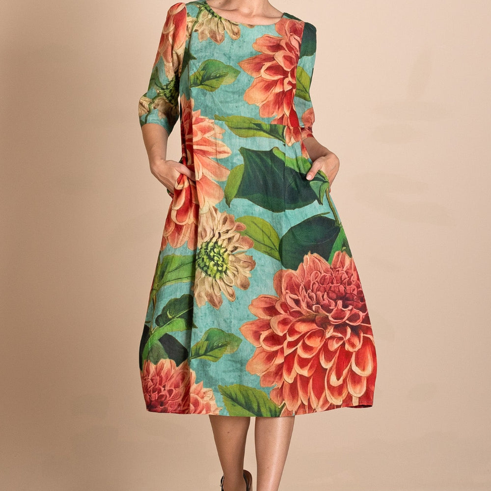 A woman models a knee  Length A-line dress with three quarter sleeves, featuring a vibrant floral print of red and orange flowers with green leaves on a turquoise blue background. She is wearing black platform sandals and posing against a solid beige studio backdrop.