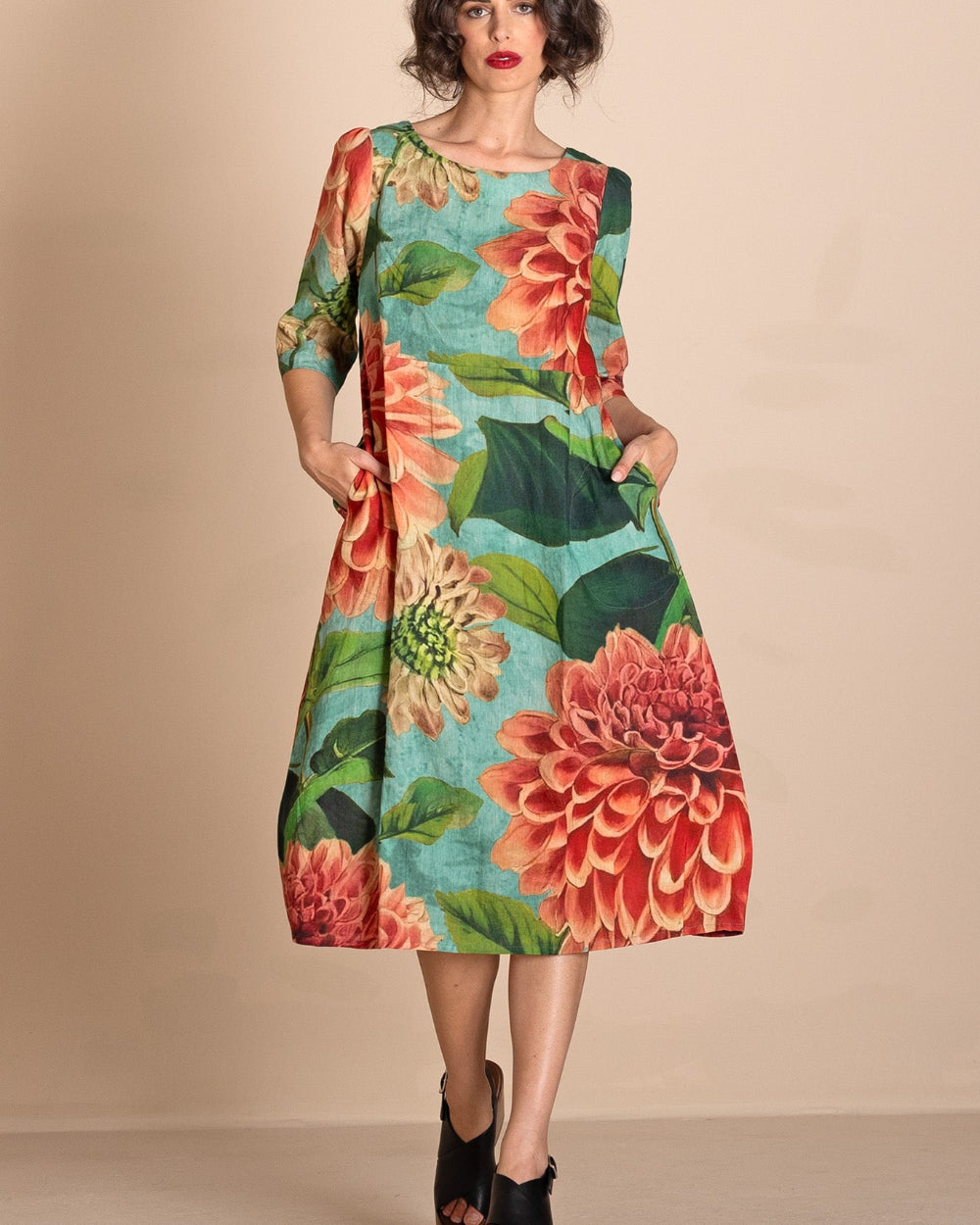 A woman models a knee  Length A-line dress with three quarter sleeves, featuring a vibrant floral print of red and orange flowers with green leaves on a turquoise blue background. She is wearing black platform sandals and posing against a solid beige studio backdrop.