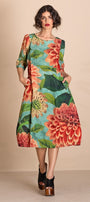 A woman models a knee  Length A-line dress with three quarter sleeves, featuring a vibrant floral print of red and orange flowers with green leaves on a turquoise blue background. She is wearing black platform sandals and posing against a solid beige studio backdrop.