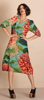 A woman models a knee length dress with a vibrant floral print featuring red and orange flowers, green leaves, and a turquoise blue background. She is posing with hands on hips against a solid beige studio backdrop.