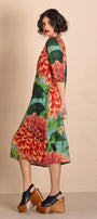 A woman modeling a knee length A-line dress with a vibrant floral print featuring red, orange, and green dahlias on a beige background.