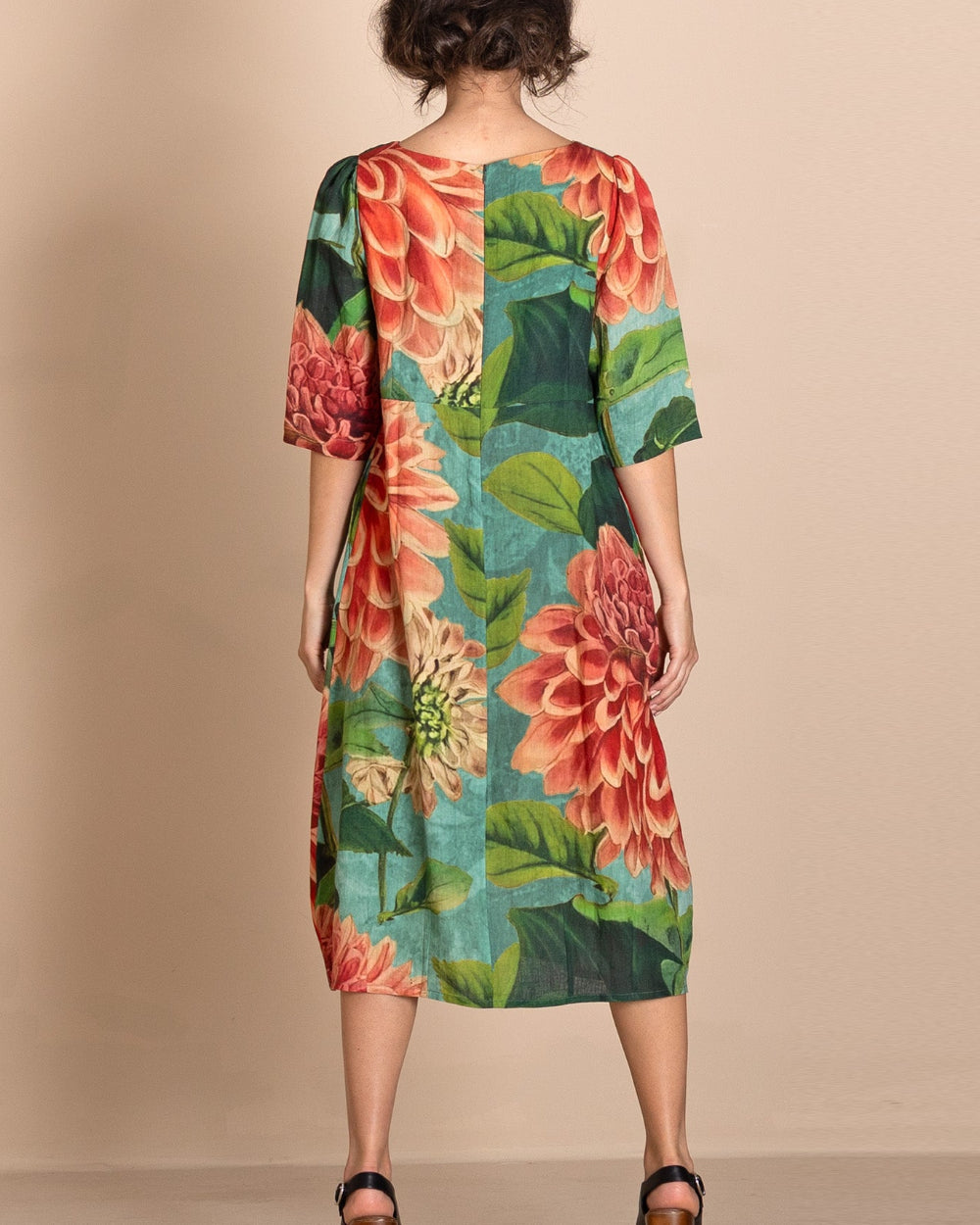 A woman viewed from the back wearing a knee length dress with a large floral print in coral, pink, green, and teal, paired with brown platform sandals, against a plain beige studio backdrop.