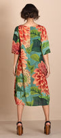 A woman viewed from the back wearing a knee length dress with a large floral print in coral, pink, green, and teal, paired with brown platform sandals, against a plain beige studio backdrop.