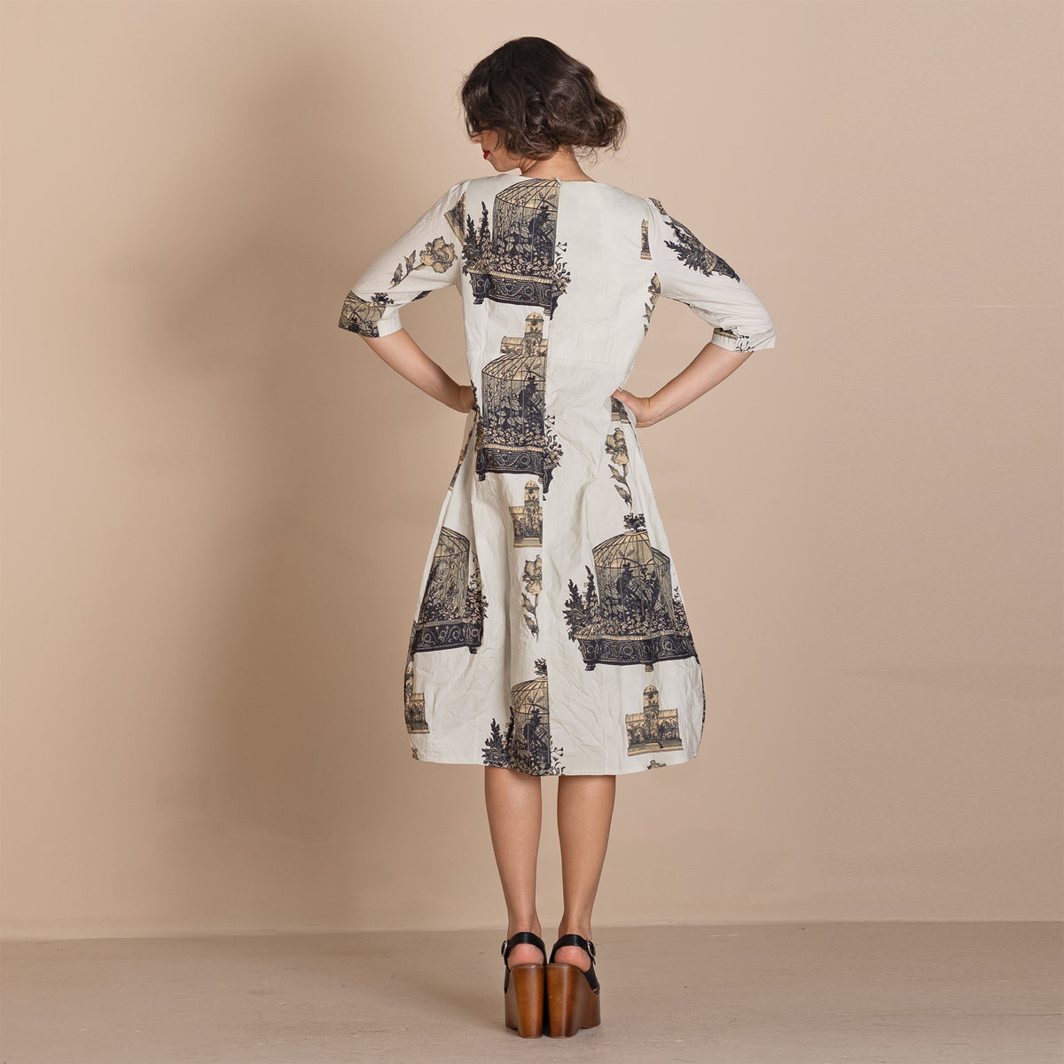  back view  wearing a knee length cream dress with a dark blue and beige architectural illustration print, paired with brown platform sandals.