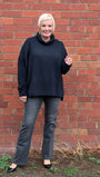 Woman wearing a dark blue sweater and gray pants standing against a brick wall.