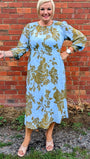Woman in a light blue dress with gold patterns standing against a brick wall.
