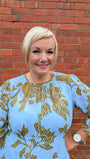 Woman wearing a blue and gold patterned dress standing against a brick wall.