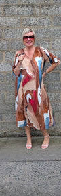 A woman wearing a beige, brown, red, and blue abstract print maxi dress and sunglasses, posing in front of a gray stone wall.