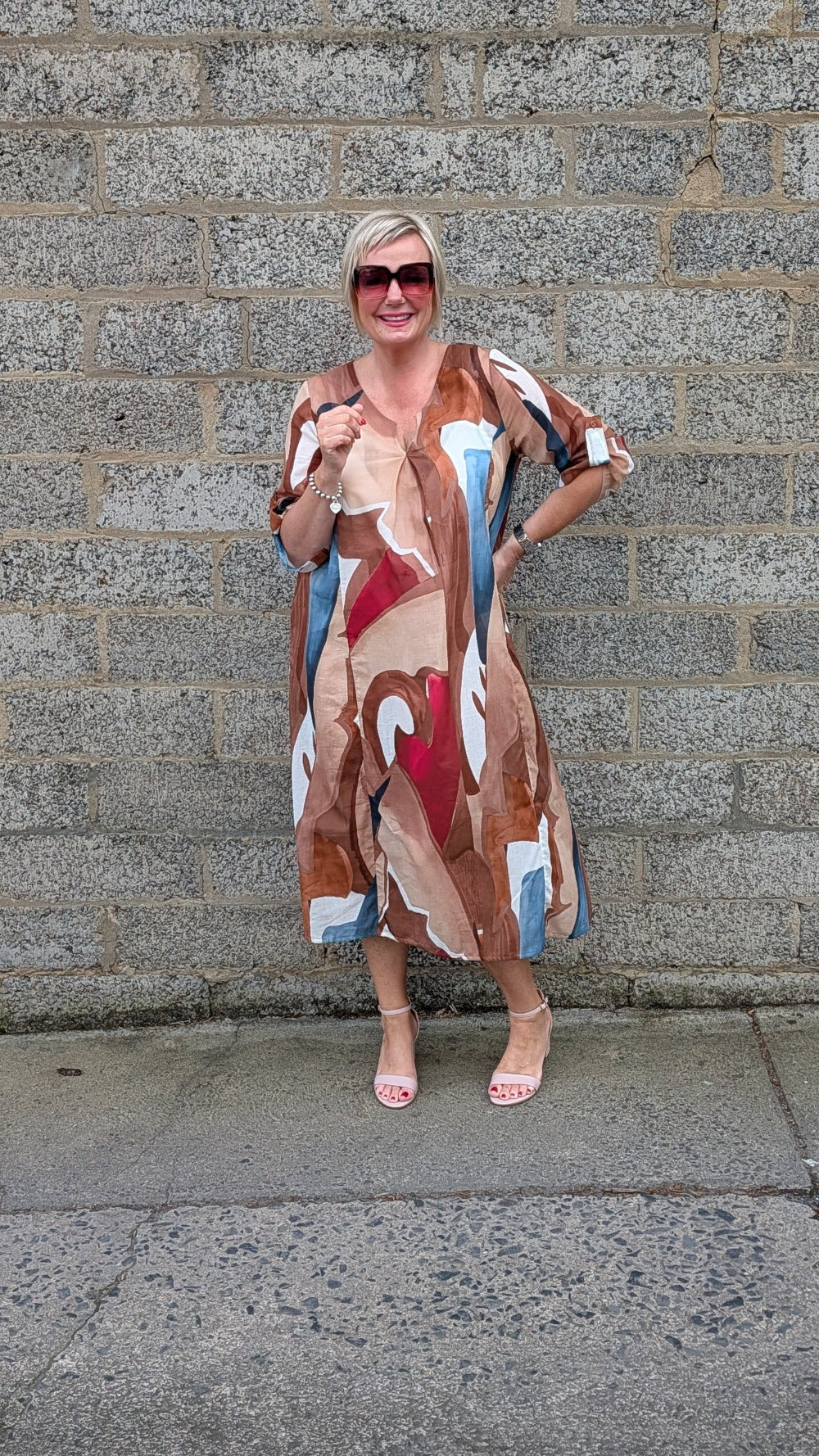 A woman wearing a beige, brown, red, and blue abstract print maxi dress and sunglasses, posing in front of a gray stone wall.
