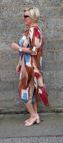 A woman wearing a long, abstract-patterned dress with brown, beige, blue, and red shapes, standing in profile against a gray stone wall.